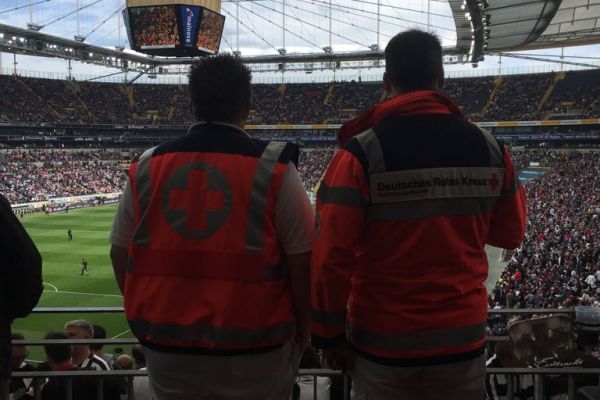 Foto: Mitarbeiter des DRK-Sanitätsdienstes blicken auf das Spielfeld im Stadion.